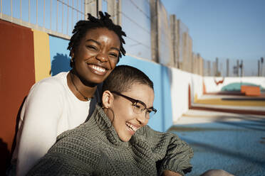 Female friends smiling while sitting at sports court - RCPF00560