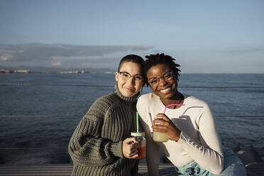 Freunde mit Saft trinken lächelnd am Meer sitzend - RCPF00568