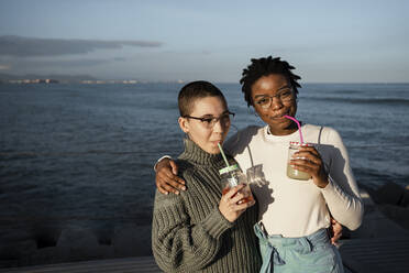 Young women drinking fruit juice while standing against sea - RCPF00571