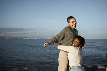 Smiling woman embracing friend standing against sea - RCPF00574