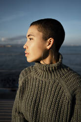 Young woman wearing turtleneck sweater looking away while sitting against sky - RCPF00579