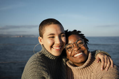 Female friends smiling while sitting against sea - RCPF00587