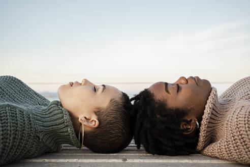 Female friends with eyes closed lying on bench against sky - RCPF00605