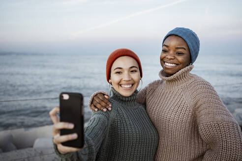 Lächelnde Freunde, die ein Selfie mit ihrem Mobiltelefon machen, während sie am Meer stehen - RCPF00609