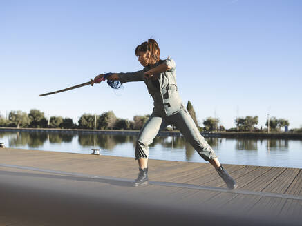 Junge Frau beim Üben von Kampfsportarten mit dem Schwert im Park am See an einem sonnigen Tag - JCCMF00902