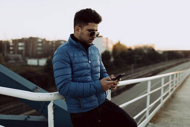 Fashionable young man using smart phone while leaning on railing against clear sky - OCAF00587