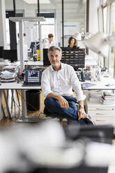 Mature businessman sitting on chair with colleague standing in background at open plan office - PESF02454