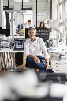 Älterer Geschäftsmann auf einem Stuhl sitzend mit einem im Hintergrund stehenden Kollegen in einem Großraumbüro - PESF02454