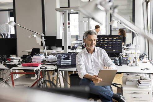 Geschäftsmann, der ein digitales Tablet benutzt, während er mit Kollegen im Hintergrund in einem Großraumbüro sitzt - PESF02455
