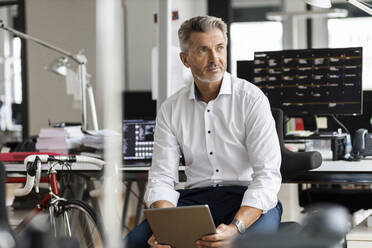 Mature businessman with digital tablet looking away while sitting at office - PESF02457