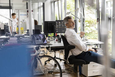 Businessman looking away while sitting on chair with colleagues in background at open plan office - PESF02461