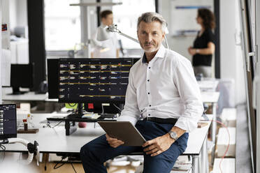 Männlicher Unternehmer mit digitalem Tablet auf dem Schreibtisch sitzend mit Kollegen im Hintergrund in einem Großraumbüro - PESF02462