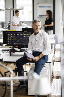 Geschäftsmann mit digitalem Tablet, der wegschaut, während er mit einem Kollegen im Hintergrund in einem Großraumbüro auf dem Schreibtisch sitzt - PESF02464
