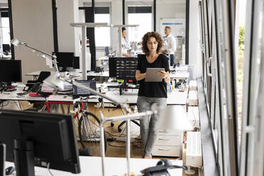 Geschäftsfrau arbeitet an einem digitalen Tablet, während sie mit einem Kollegen im Hintergrund in einem Großraumbüro steht - PESF02468