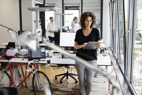 Unternehmerin arbeitet an einem digitalen Tablet, während sie mit einem Kollegen im Hintergrund in einem Großraumbüro steht - PESF02470