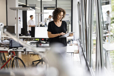Geschäftsfrau, die ein digitales Tablet benutzt, während sie mit einem Kollegen im Hintergrund in einem Großraumbüro arbeitet - PESF02471