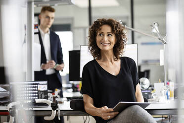 Smiling businesswoman using digital tablet while sitting with colleague in background at open plan office - PESF02477