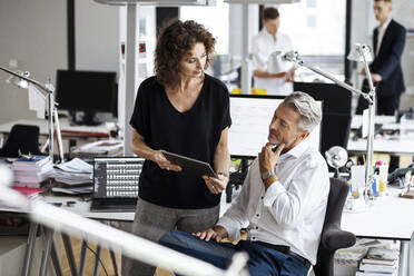 Ältere Geschäftsleute, die ein digitales Tablet benutzen, während sie mit Kollegen im Hintergrund in einem Großraumbüro arbeiten - PESF02480