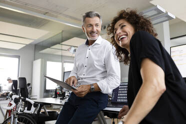 Lächelnder Geschäftsmann mit digitalem Tablet, der einen fröhlichen Kollegen ansieht, während er in einem Großraumbüro steht - PESF02489