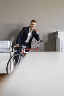 Selbstbewusster Geschäftsmann mit Hand am Kinn, der sich auf ein Fahrrad stützt, während er im Büro steht - PESF02499