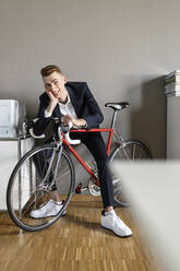 Lächelnder Geschäftsmann sitzt mit Hand am Kinn auf einem Fahrrad im Büro - PESF02501