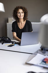 Nachdenkliche Geschäftsfrau lächelt, während sie im Büro sitzt - PESF02503