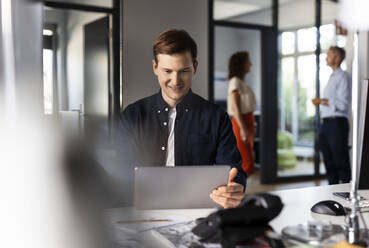 Smiling businessman using digital tablet while sitting with colleagues in background at open plan office - PESF02505