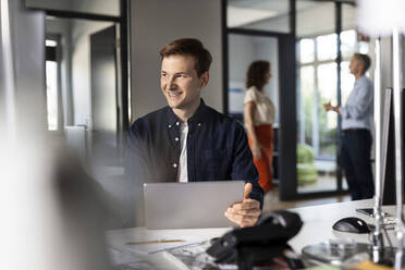 Smiling businessman looking away while sitting with colleagues in background at open plan office - PESF02506