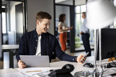 Young businessman using mobile phone while sitting with colleague in background at open plan office - PESF02507