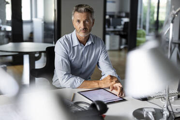Älterer selbstbewusster Geschäftsmann mit digitalem Tablet, der im Büro sitzt und starrt - PESF02516