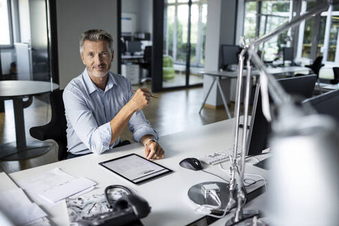 Älterer Unternehmer mit digitalem Tablet, der lächelnd im Büro sitzt - PESF02518