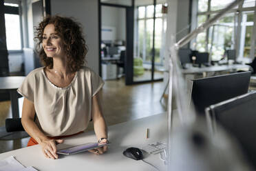 Lächelnde reife Frau mit digitalem Tablet, die wegschaut, während sie in einem Großraumbüro sitzt - PESF02519