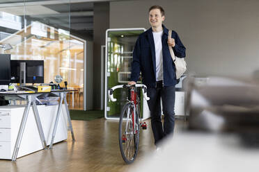 Lächelnder Geschäftsmann mit Tasche und Fahrrad, der nach der Arbeit aus dem Büro geht - PESF02521