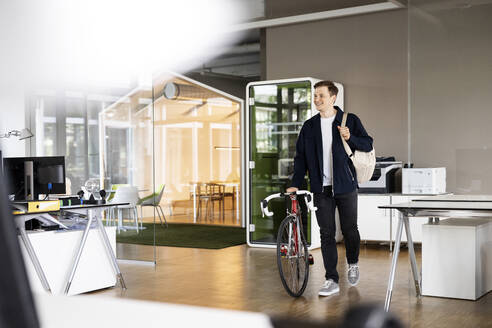 Smiling businessman walking with bicycle at open plan office - PESF02524