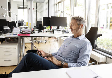 Businessman using digital tablet while relaxing on chair at open plan office - PESF02533