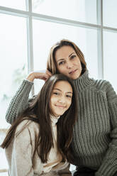 Smiling mother and daughter sitting against glass window at home - RCPF00633