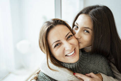 Daughter kissing happy mother by window at home stock photo