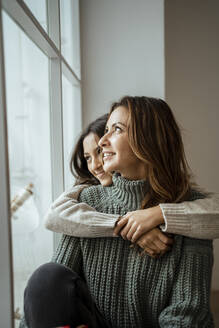 Happy mother and daughter looking through window while day dreaming together at home - RCPF00639