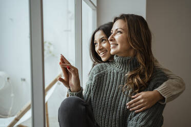 Happy mother and daughter looking through window while sitting at home - RCPF00641