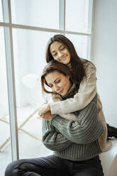 Mother and daughter embracing each other with eyes closed while sitting on window seat at home - RCPF00644