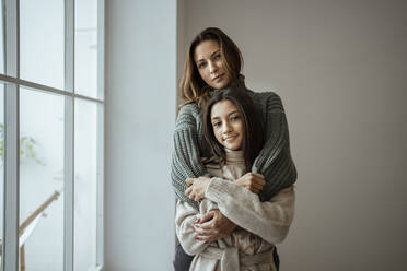 Mutter und Tochter umarmen sich gegenseitig, während sie zu Hause an der Wand stehen - RCPF00647