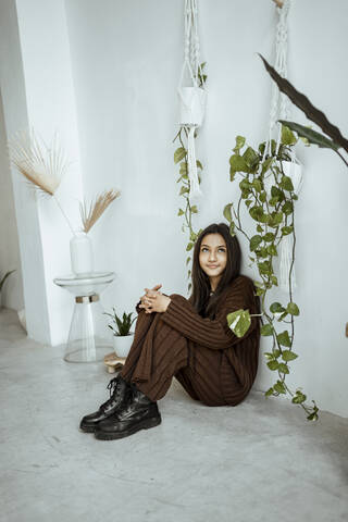 Smiling girl looking up while sitting by creeper plant against white wall at home stock photo