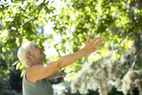 Carefree senior woman with arms outstretched under summer trees - CAIF30082