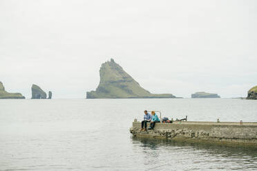 Travelers sitting on breakwater on cloudy day - CAVF91765