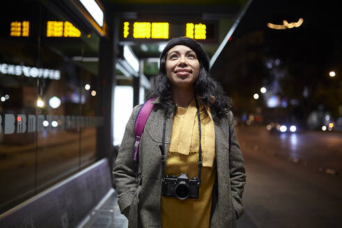 Woman standing with a camera waiting at the bus station at night - CAVF91793