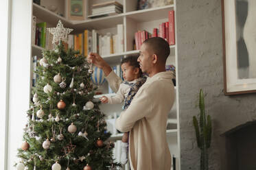 Father and baby daughter decorating Christmas tree in living room - CAIF30113