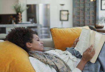 Serene woman reading book on living room sofa - CAIF30119