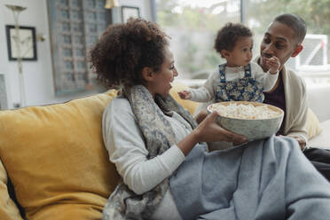 Glückliche Eltern und ihre kleine Tochter beim Fernsehen mit Popcorn auf dem Sofa - CAIF30127