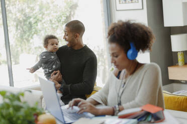 Vater hält seine kleine Tochter hinter der arbeitenden Mutter am Laptop - CAIF30150