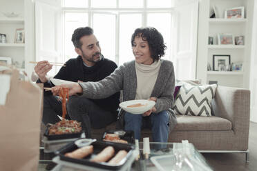 Glückliches Paar genießt Essen zum Mitnehmen mit Stäbchen auf dem Wohnzimmersofa - CAIF30173